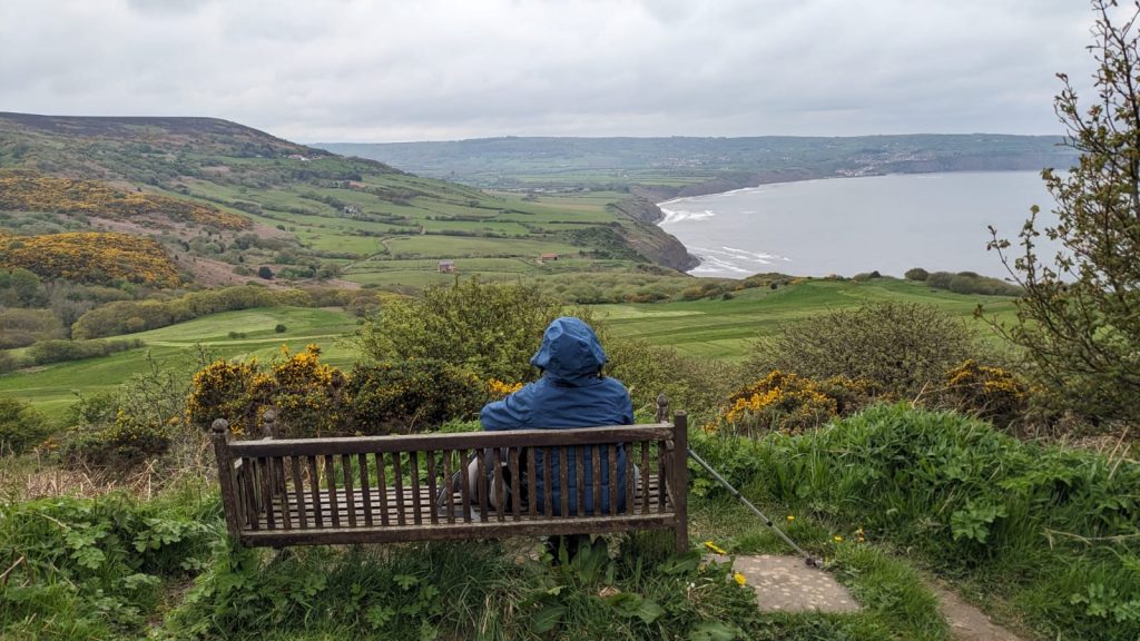 A lonely hiker is sitting on a bench looking over a lush green valley toward Robin Hood's Bay.