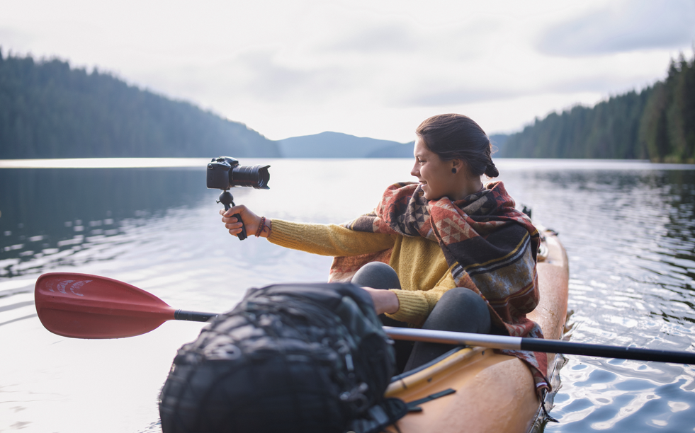 A vlogger sitting in a kayak on a calm lake surrounded by mountains and forrest. Content creation can be an amazing experience.