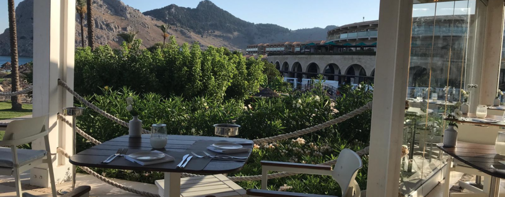 View from a restaurant in Rhodes, Greece. An empty table and bushes. Mountains in the distance against clear blue sky.