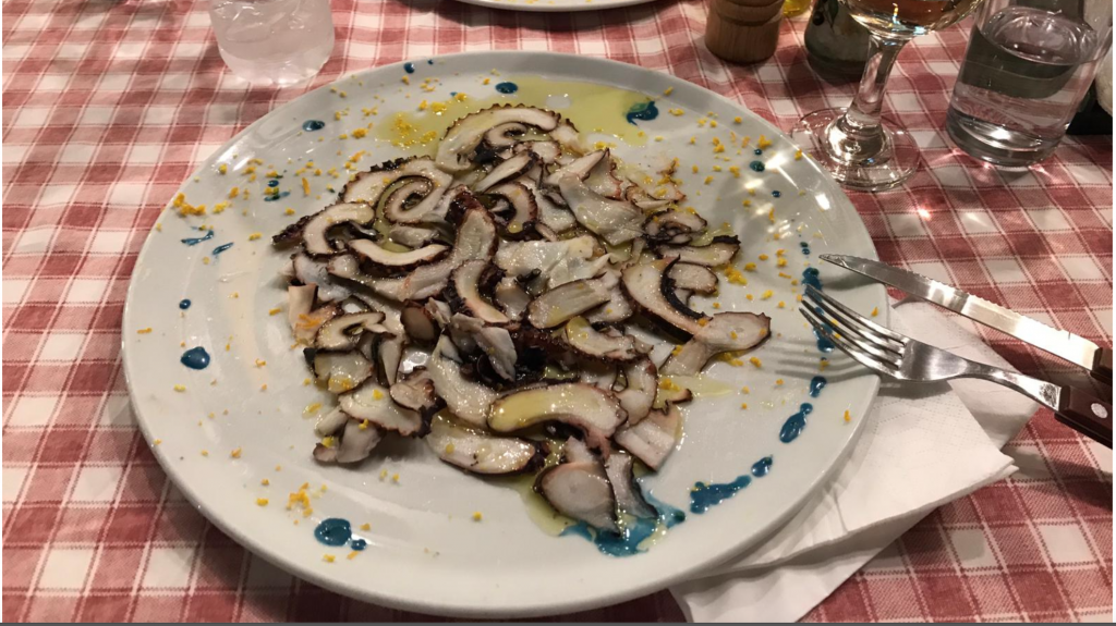 Pictured a plate of thinly sliced octopus carpaccio started on top of checkered tablecloth.
