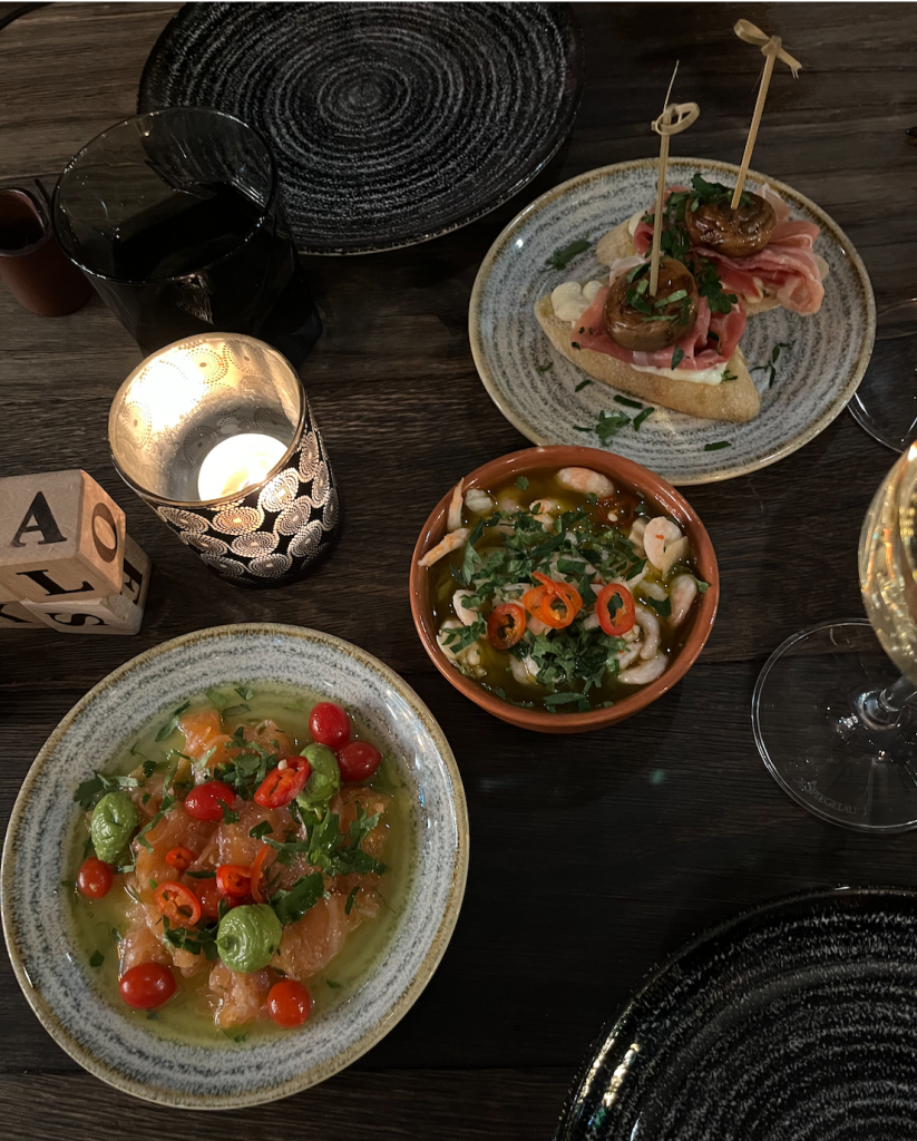 Pictured three small plates of tapas on top of brown wooden table, including salmon ceviche, shrimp in garlic sauce and serrano ham and mushroom served on bread.