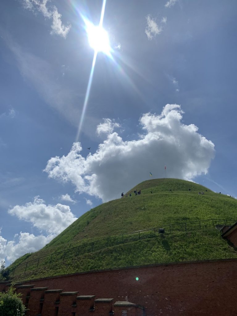 Sunshine and clouds. A big green mound with people walking up there. On the top there is a flags of Poland and Ukraine.