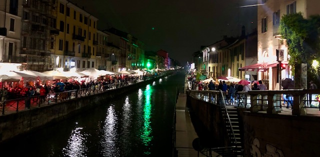 Navigli Canal in the evening. The canal is full of restaurants, lots of atmospheric lights everywhere, and the air is filled with people talking and the sound of rain.