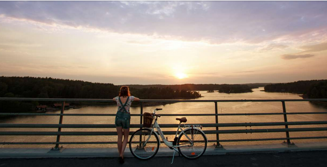 Tyttö seisoo sillalla polkupyörän vieressä ja katselee merelle auringonlaskua.
