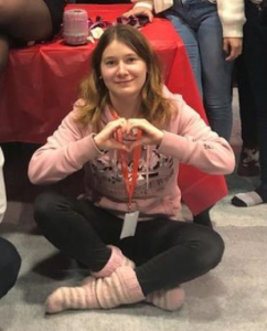 An image of the person the text is about. She is sitting on the floor and making a shape of a heart with her fingers.