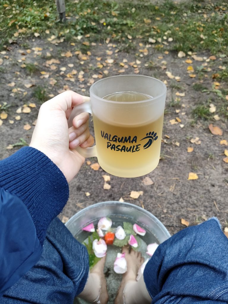 Soaking feet in hot water with some herbs and rose petals while drinking herbal tea in a glass cup of Valguma Pasaule after the barefoot trail