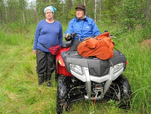 Kaksi iäkästä henkilöä metsän reunalla. Toinen istuu mönkijän ohjaimissa ja toinen seisoo sen vieressä.