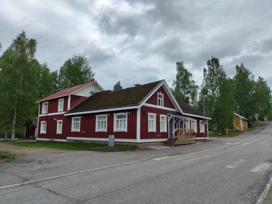 A red wooden building with white decorative trim stands beside the road. The building is surrounded by trees, and the sky is cloudy.