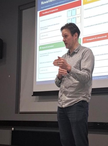 Reko Silvenius gestures while presenting in front of a projected workshop framework with sections on safety, sales and responsibilities, and partners and stakeholders.