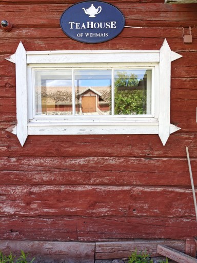 White-trimmed window set in a red log wall. Above the window is a sign reading “TeaHouse of Wehmais,” and the glass reflects a yard building and surrounding greenery.