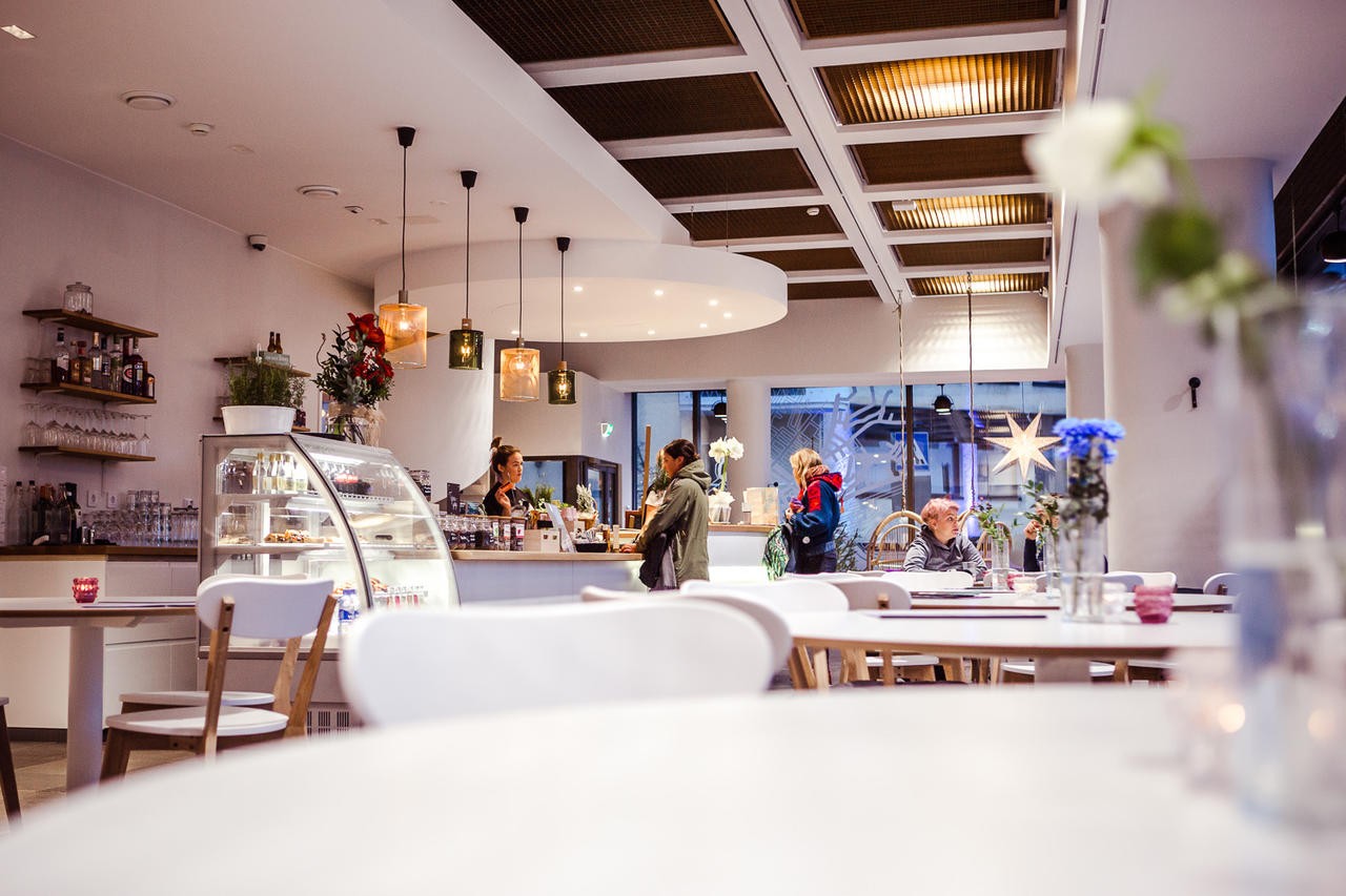 Cafe Hostel Kodi restaurant interior with white tables, a service counter, and customers in a cozy, modern café setting.