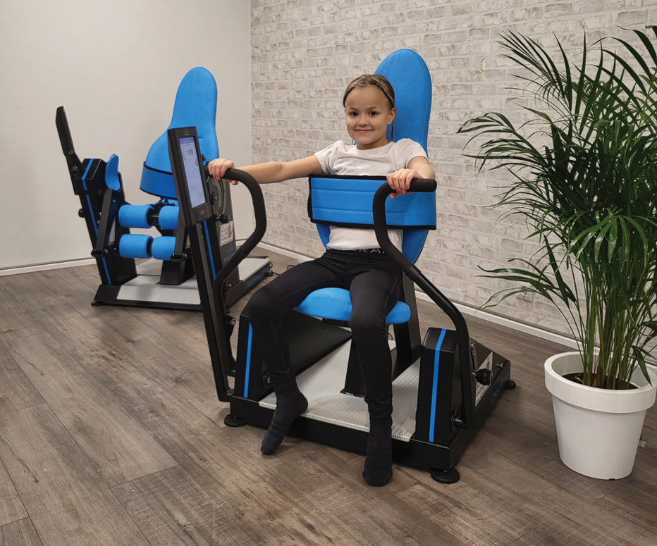 A smiling child is seated on a gym machine, holding the machine's handles. The equipment is either a chest press or a back extension machine and features blue padding.