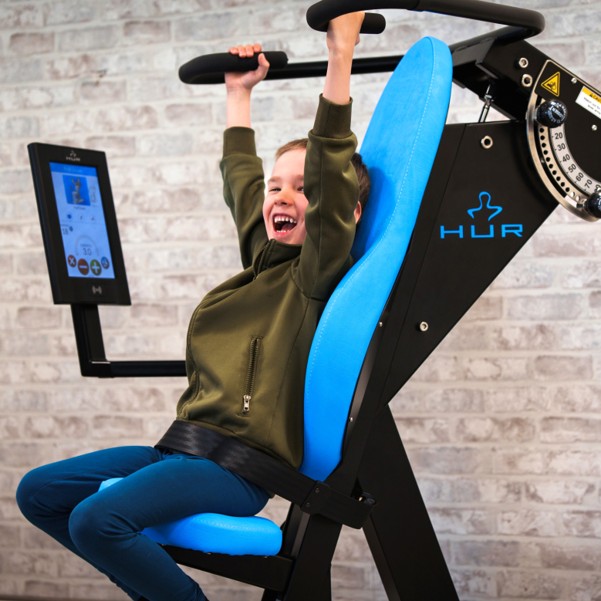 A smiling child is seated on a strength-training machine, holding the overhead handles while performing an exercise.