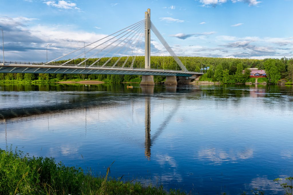 Lumberjack's Candle Bridge in the summer at Rovaniemi's center. Clear blue water, green vegetation, and light blue sky. Restaurant Valdemari can be seen in the background. 