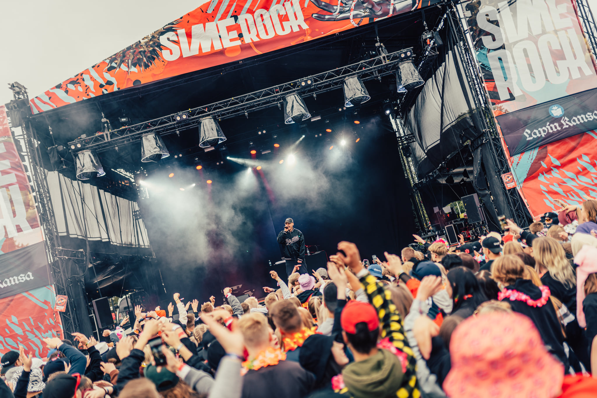 Sime rock summer festival in Rovaniemi. A crowd of people in the audience in front of a stage shot from behind. Many of them have their hands in the air. On the stage, there is a rapper performing.