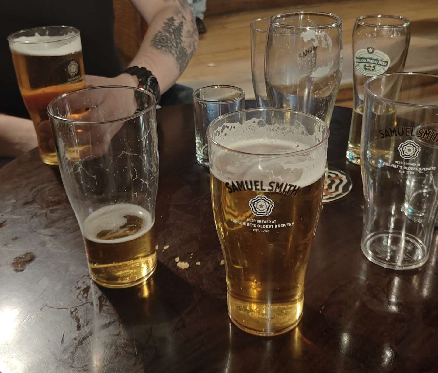 Tumman puupöydän äärellä useita puolityhjiä ja tyhjiä olutlaseja, joissa Samuel Smithin logo; taustalla henkilö mustassa paidassa ja tatuoinnilla varustetulla käsivarrella pitelee tuoppia.