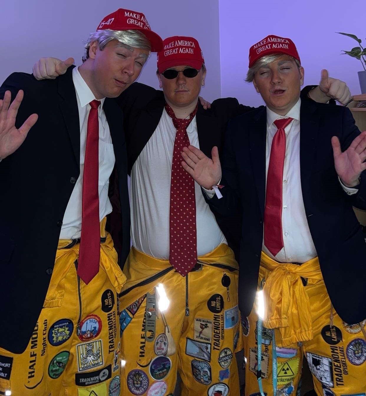 Kolme henkilöä pukeutuneena punaisiin 'Make America Great Again' -lippiksiin, tummaan pikkutakkiin, valkoiseen paitaan ja keltaisiin opiskelijahaalareihin, poseeraavat leikillisesti sisätiloissa.