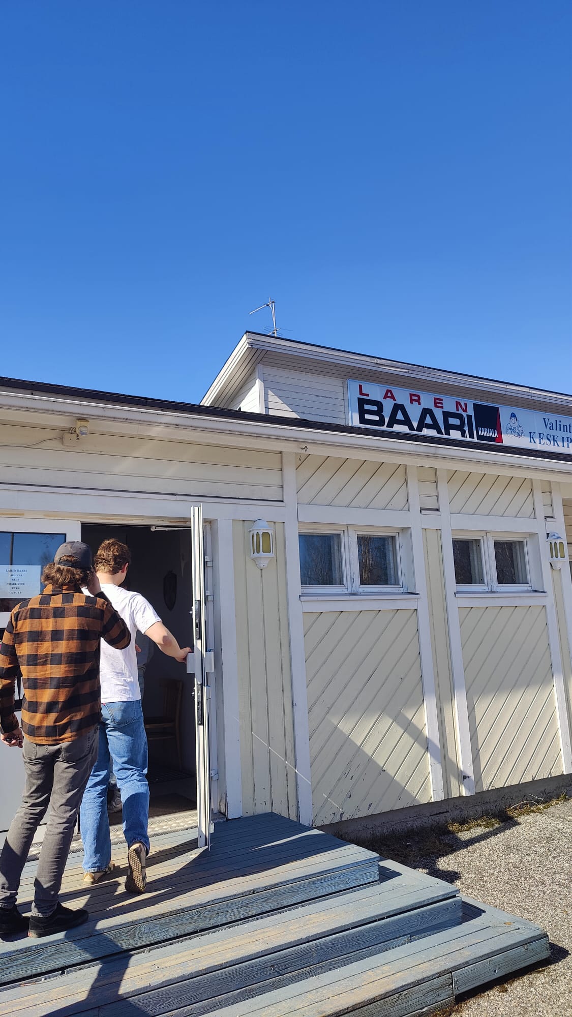 Paavo ja Fabian kävelee valkoisen puurakennuksen sisäänkäynnille, jossa yläpuolella lukee "Laren Baari", aurinkoisena ja pilvettömänä päivänä.