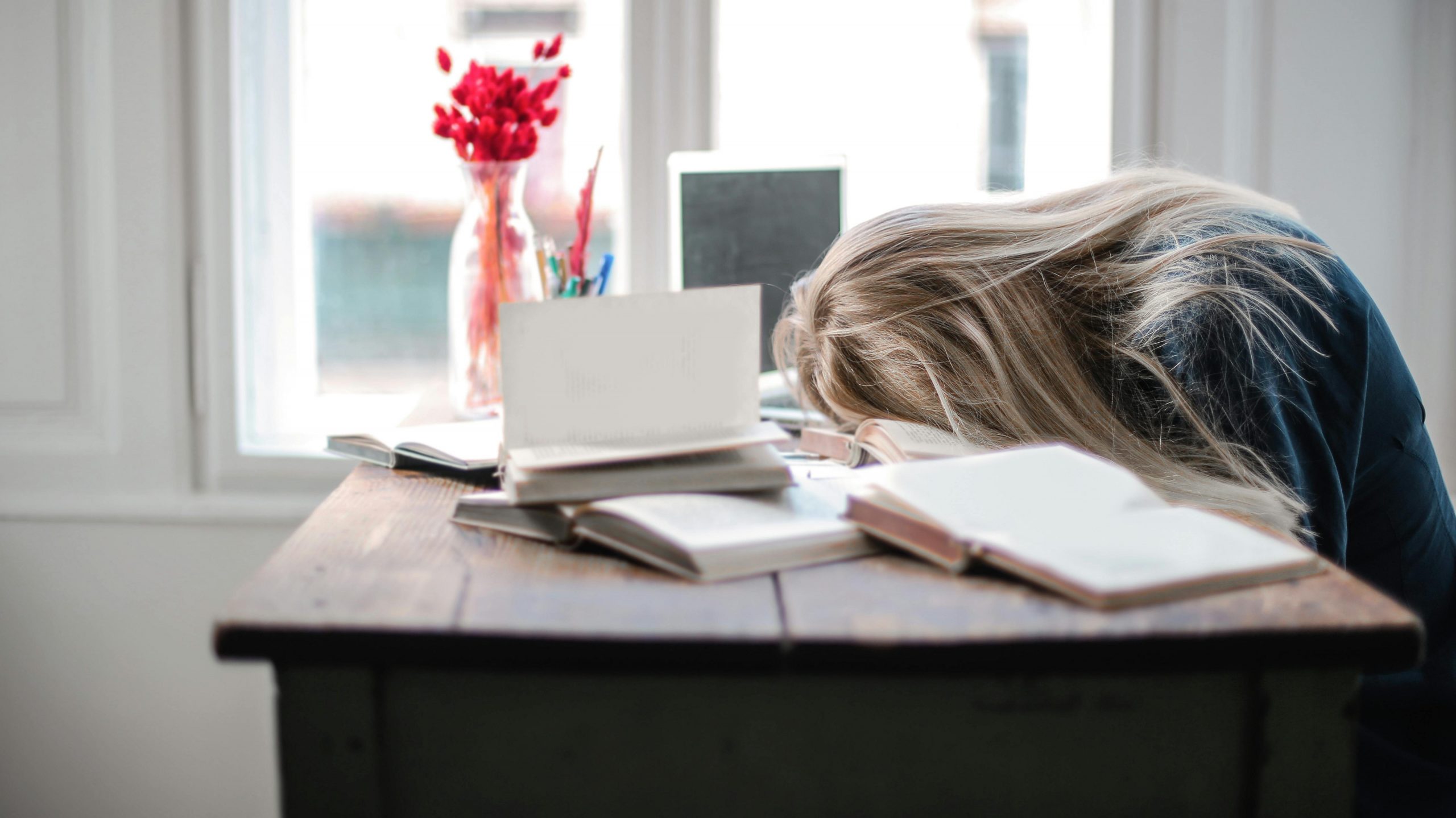 Väsynyt ja stressaantunut opiskelija nojaa päätään kirjojen päälle työpöydällä. Taustalla on ikkuna. 
