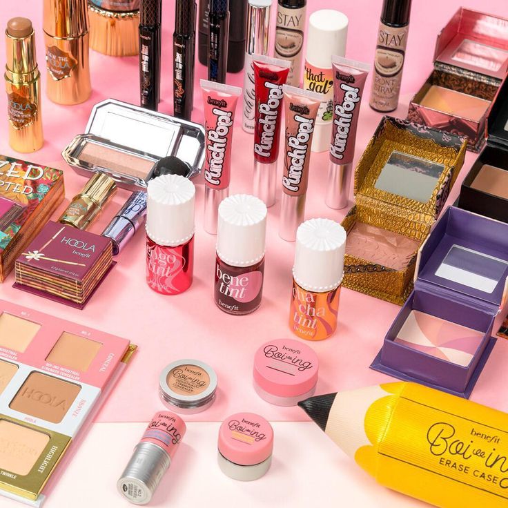 Benefit Cosmeticsin meikkikokoelma vaaleanpunaisella taustalla: huulituotteita, poskipunia, highlightereita, peitevoiteita, ripsivärejä ja sävyttävää poskipunaa brändin tunnistettavissa pinkeissä ja retrohenkisissä pakkauksissa. Tuotteet on aseteltu selkeään, inspiroivaan flat lay -kuvaan, joka korostaa Benefit-brändin värikästä ja leikkisää estetiikkaa.