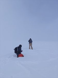 Kaksi vapaalaskijaa huonossa näkyvyydessä (whiteout) huipun lähellä. Toinen laskijoista polvillaan lumessa.
