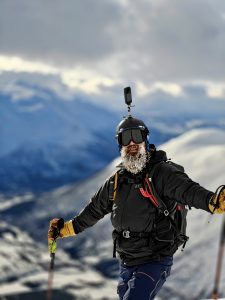 Lähikuva iloisesta vapaalaskijasta SØrfjelltindenin huipulla. Parta ja kulmakarvat ovat huurteessa, kypärässä actionkamera.