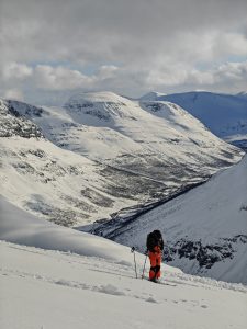 Vapaalaskija seisoo rinteellä katsellen alas kohti laaksoa Lyngen-vuoristossa. Laskija oransseissa housuissa ja mustassa repussa.