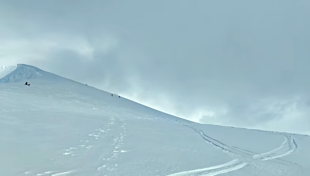 Etäällä näkyviä vapaalaskijoita nousemassa jyrkkää lumista rinnettä SØrfjelltindenillä Lyngenissä. Aikaisemmat laskujäljet näkyvissä.