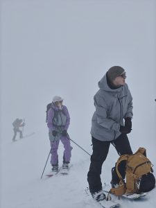 Kaksi vapaalaskijaa sumuisessa ja heikossa näkyvyydessä nousun aikana. Toisella on päällään harmaa takki ja toisella violetti.