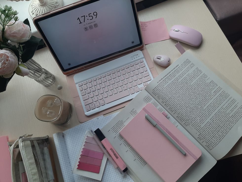 Tyylikäs vaaleanpunasävyinen työpöytäasetelma, jossa on avoin kirja, muistiinpanovälineitä, tabletilla varustettu näppäimistö, vaaleanpunaiset hiiret, värimallit, kukkakimppu ja lasillinen jääkahvia.